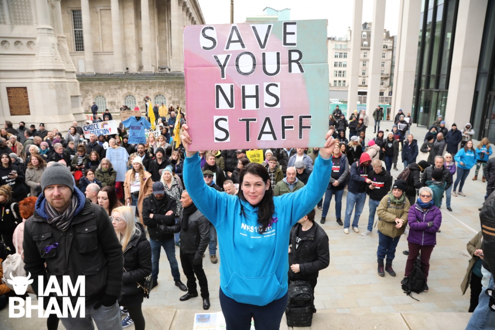 Protester wearing a blue NHS100k hoodie, holds up a placard
